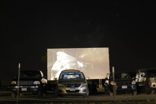 A drive-in movie screen displays a film under a starry night sky, with parked cars in the foreground.