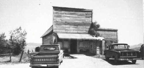 Black and white photo of an old wooden building with two vintage Chevrolet trucks parked outside.