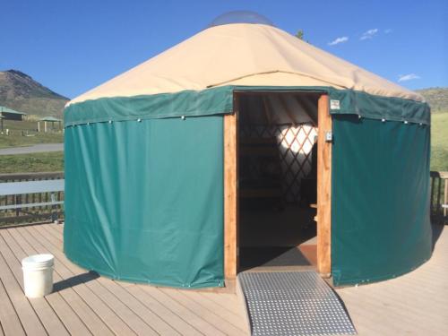 A green yurt with a tan roof, set on a wooden deck, surrounded by grassy hills and blue sky.