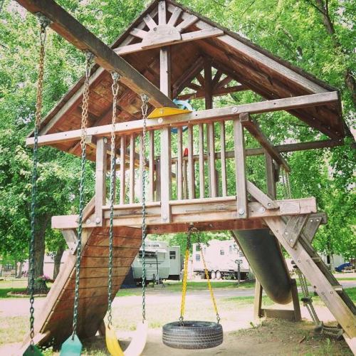 Wooden playground structure with a slide, swings, and a tire swing, surrounded by trees and trailers in the background.