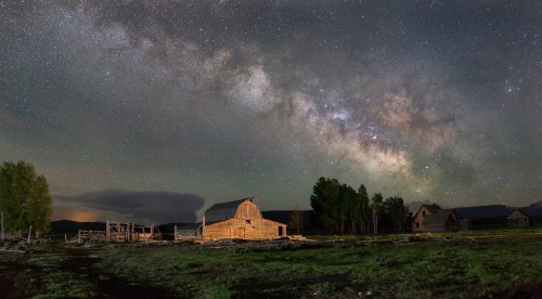 A rustic barn under a starry night sky filled with the Milky Way, surrounded by trees and open fields.