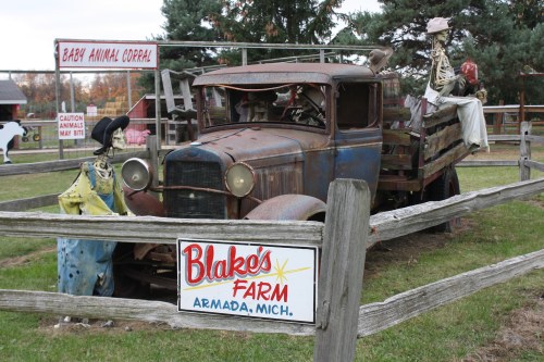 A vintage truck with skeletons sits in a farmyard, surrounded by a wooden fence and a sign for Blake's Farm.