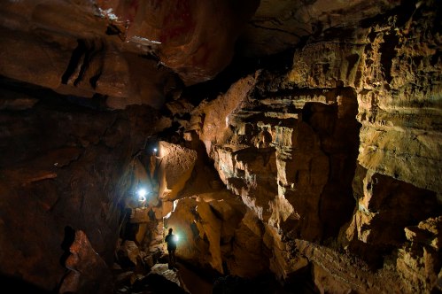 A person stands in a dimly lit cave, surrounded by rugged rock formations and illuminated by small lights.