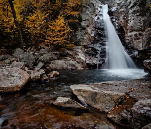 A serene waterfall cascades over rocky cliffs, surrounded by autumn foliage and a tranquil pool below.