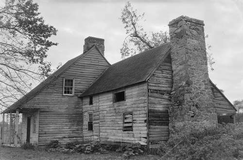 Black and white photo of an old, weathered wooden house with a stone chimney and overgrown vegetation.