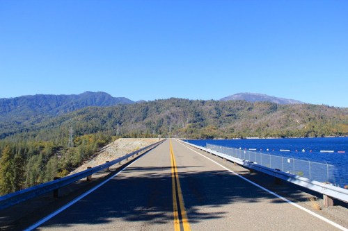 A straight road stretches towards mountains, bordered by a lake and clear blue sky.