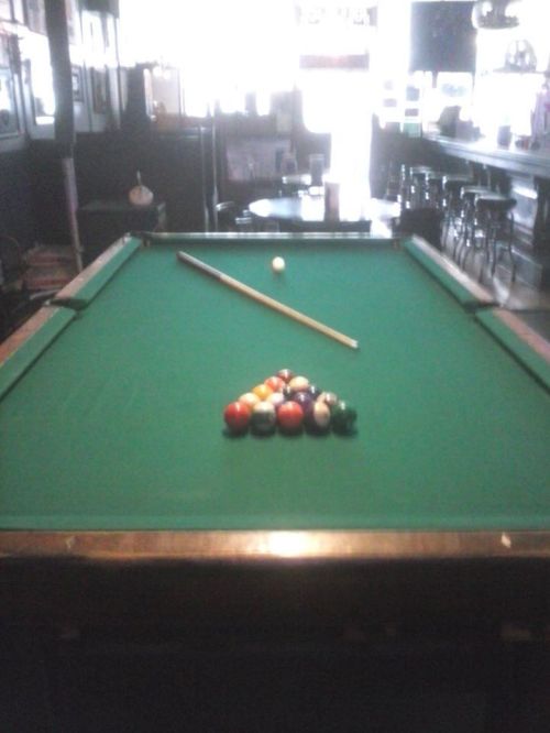 A pool table with a triangle of colored balls, a cue stick, and a single white ball in a bar setting.