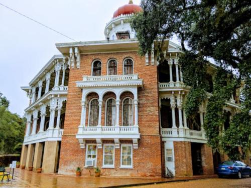 A historic brick building with ornate white balconies and a red dome, surrounded by greenery on a rainy day.