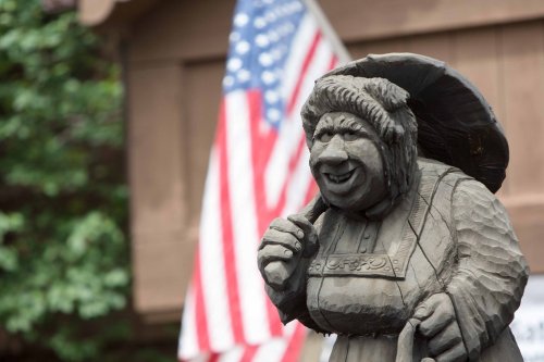 A carved wooden statue of a smiling woman holding an umbrella, with an American flag in the background.