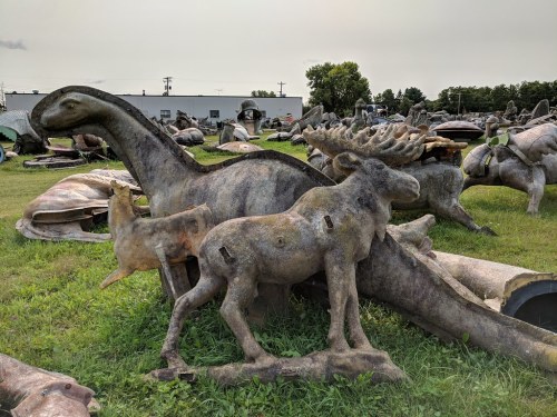 A collection of large, weathered animal sculptures, including a dinosaur, moose, and other creatures, in a grassy field.