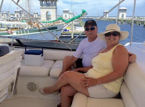 A couple relaxes on a boat, smiling and enjoying the view of a marina with boats in the background.