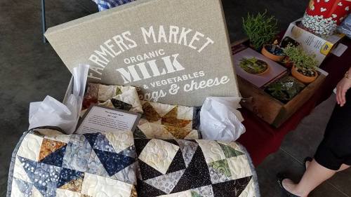 A farmers market display featuring organic milk, vegetables, and a quilted tablecloth with a variety of plants in the background.