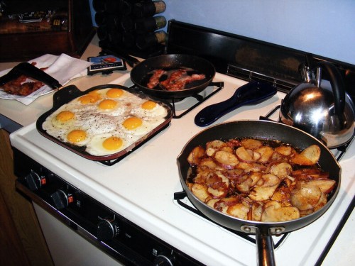 A kitchen stove with two frying pans: one with fried eggs and bacon, the other with sautéed potatoes.