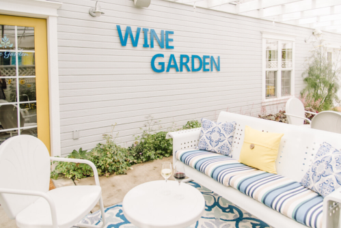 A cozy outdoor wine garden with a white sofa, colorful cushions, and a sign that reads "WINE GARDEN."