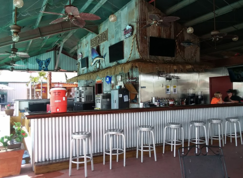 A rustic bar with metal stools, tropical decor, and a vibrant atmosphere, featuring fans and a variety of drinks.