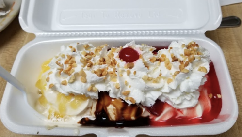 A takeout container filled with ice cream topped with whipped cream, nuts, a cherry, and colorful syrups.