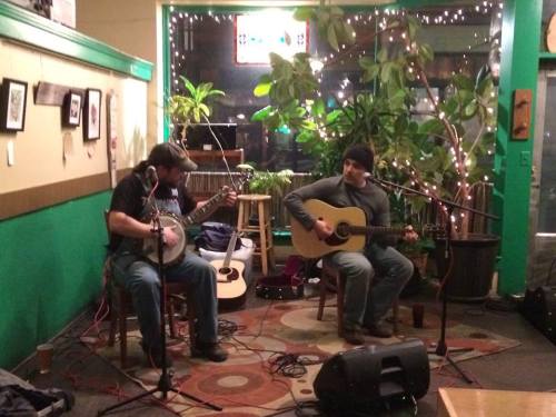 Two musicians perform on stage in a cozy venue, surrounded by plants and warm lighting. One plays banjo, the other guitar.