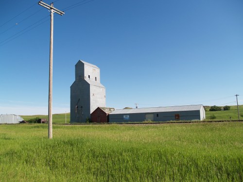 A tall, gray grain elevator stands beside a low metal building, surrounded by green grass under a clear blue sky.