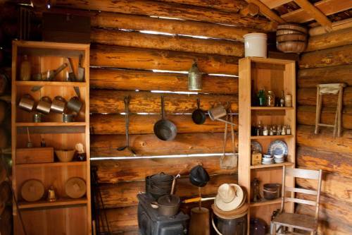 Interior of a rustic cabin with wooden walls, shelves of vintage kitchenware, and various tools hanging.