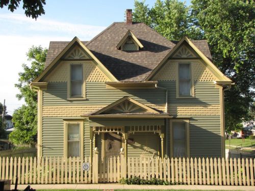 A green Victorian-style house with a peaked roof, decorative trim, and a white picket fence in front.