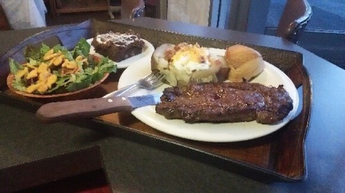 A plate with a grilled steak, baked potato topped with sour cream, salad, and a roll on a wooden tray.