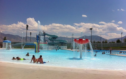A sunny water park scene with children swimming, water slides, and splash features against a mountain backdrop.
