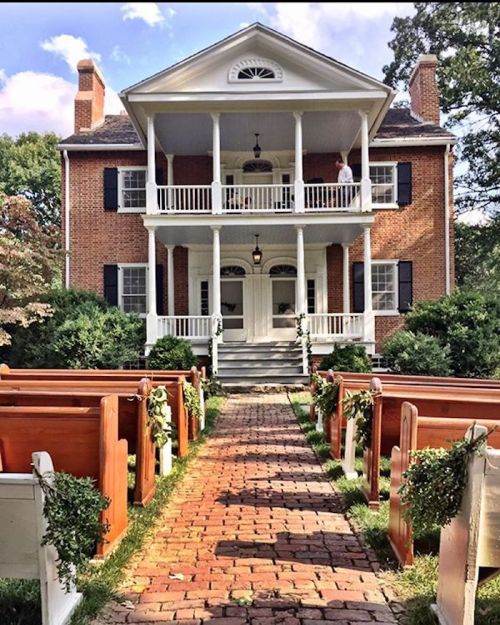 A charming brick house with a porch, flanked by wooden benches adorned with greenery, leading to a wedding setup.