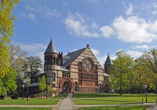 Historic building with ornate architecture, towers, and large windows, surrounded by green trees and a clear blue sky.