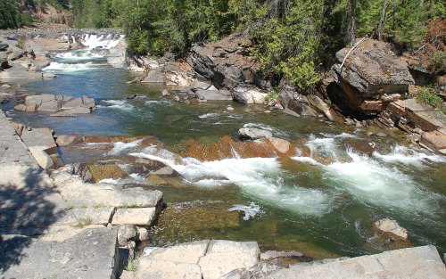 A serene river flows over rocky terrain, surrounded by lush green trees and cascading waterfalls in the background.
