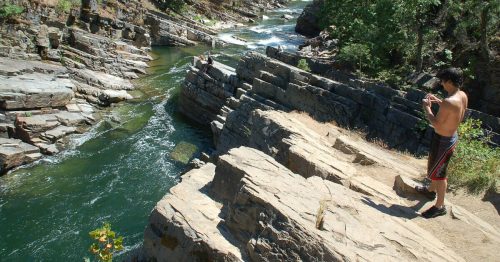 A shirtless person stands on rocky terrain by a river, surrounded by trees and steep cliffs.