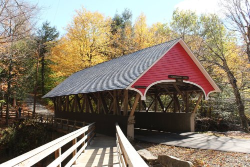 A pink covered bridge surrounded by autumn trees, with a wooden walkway leading to it.