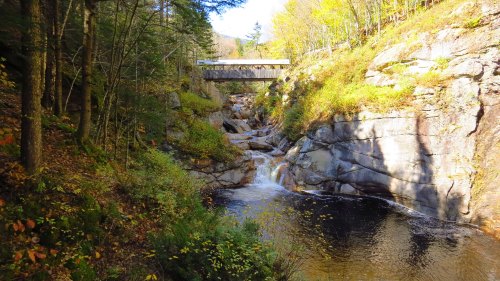 A serene river flows through rocky terrain, surrounded by autumn foliage and a bridge overhead.