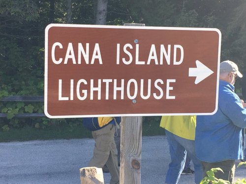 Brown sign pointing to Cana Island Lighthouse with a person in blue clothing nearby. Green foliage in the background.