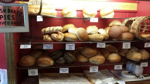 A display of various freshly baked breads on shelves, including rolls and loaves, with price tags visible.