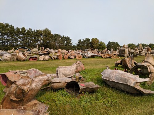 A field filled with various large, weathered sculptures and art pieces, surrounded by trees and grass.