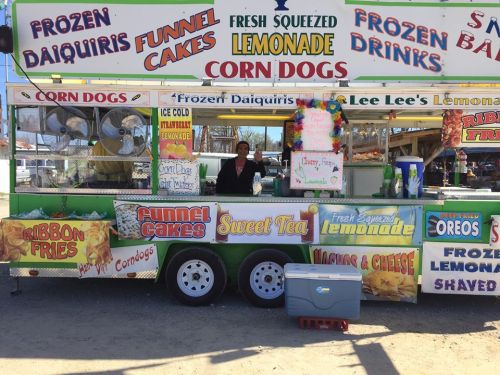 A colorful food truck offering frozen daiquiris, funnel cakes, lemonade, corn dogs, and other treats at a fair.