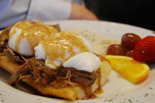 A plate of eggs benedict with pulled meat, hollandaise sauce, and a side of fruit including grapes, orange, and strawberry.