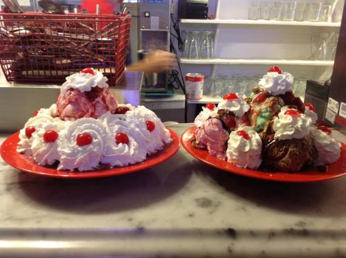 Two large ice cream sundaes on red plates, topped with whipped cream and cherries, featuring various flavors of ice cream.