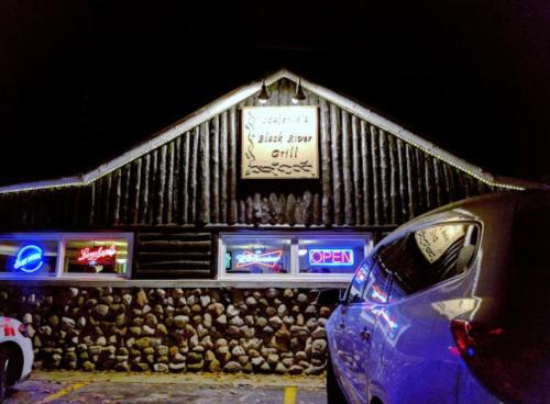 A rustic restaurant exterior at night, featuring a sign that reads "Black River Grill" and colorful neon lights.