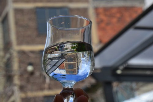 A glass of water reflecting a building upside down, with a blurred background of urban architecture.