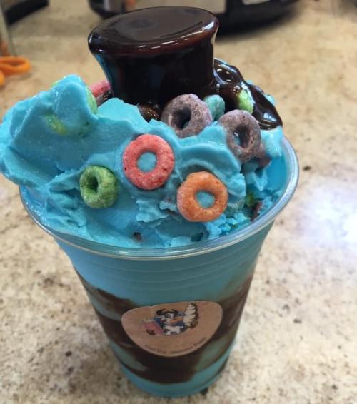 A colorful dessert in a cup, featuring blue whipped cream, chocolate sauce, and fruity cereal toppings.