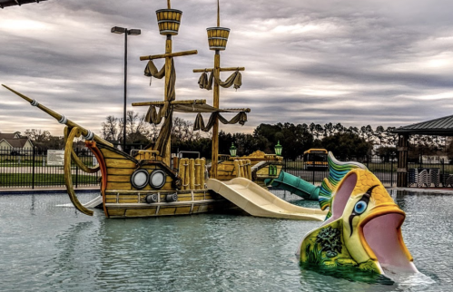 A pirate ship playground with a water slide and a colorful fish sculpture in a pool area.