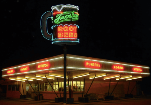 Neon sign for Frodo's Root Beer, featuring a large mug, with a retro diner serving burgers and hot dogs at night.
