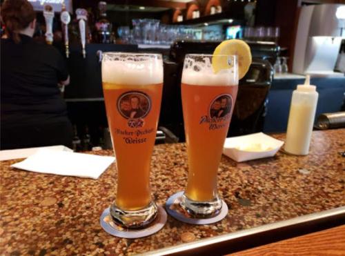 Two glasses of beer with lemon slices on a bar counter, with a blurred background of taps and bottles.