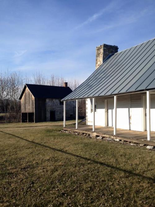 A grassy area with two buildings: a stone house with a chimney and a wooden barn in the background under a clear sky.