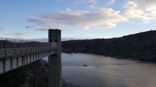 A tall structure beside a calm lake, with hills and clouds in the background, under a clear blue sky.