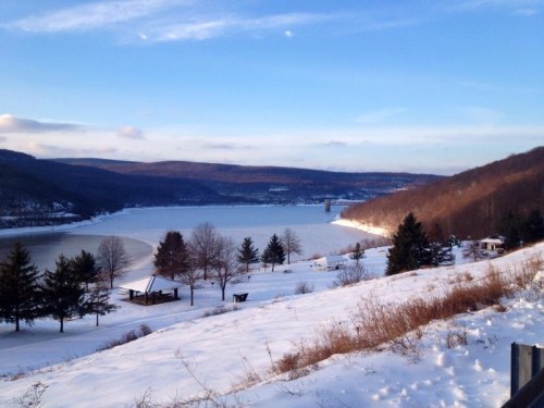 A snowy landscape with a river, trees, and distant hills under a clear blue sky.