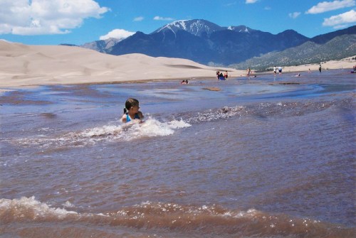 A child plays in a shallow stream at a sandy desert, with mountains and blue sky in the background.