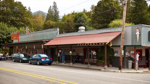 A small town street featuring shops with metal roofs, parked cars, and people outside enjoying the day.