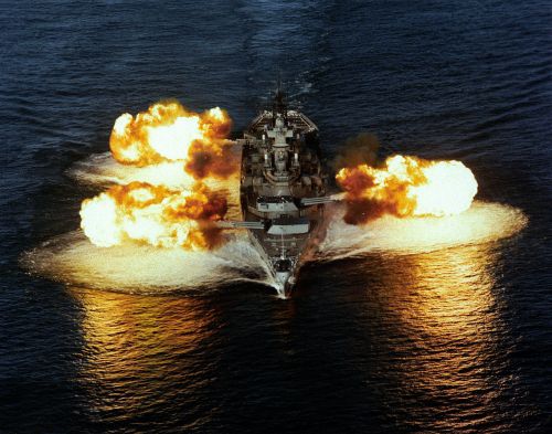 A battleship fires its cannons, creating large explosions in the water, with flames and smoke against a blue ocean backdrop.